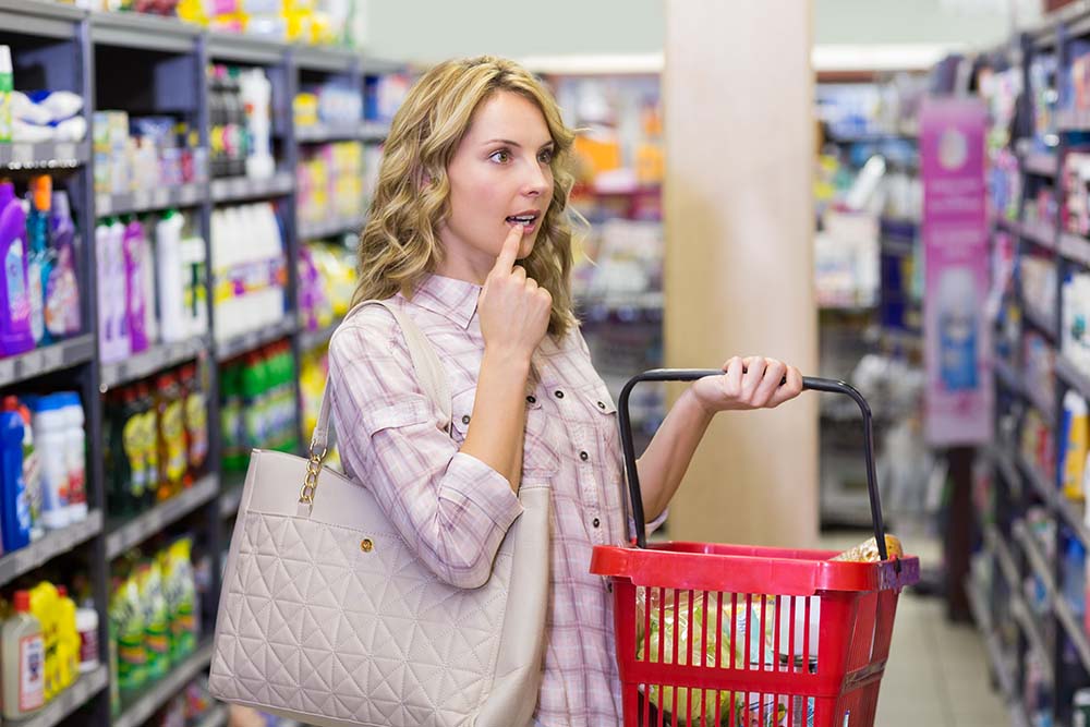 Frau mit Einkaufstasche blickt nachdenklich auf das Supermarktregal