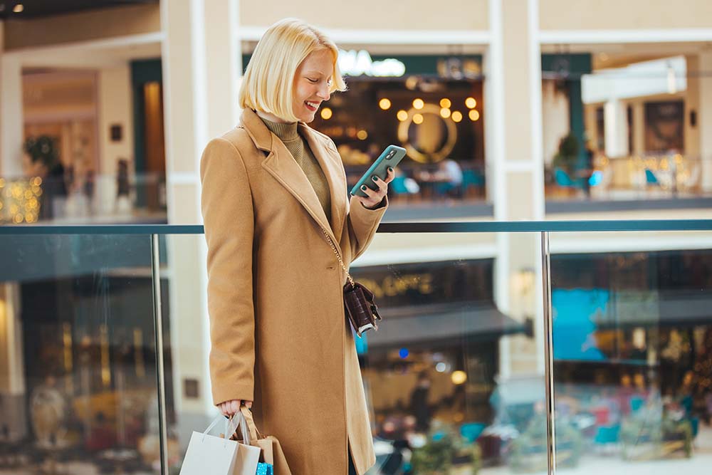 Junge Frau mit Einkaufstüten nutzt Smartphone, während sie im Geschäft steht (Bulgarien)