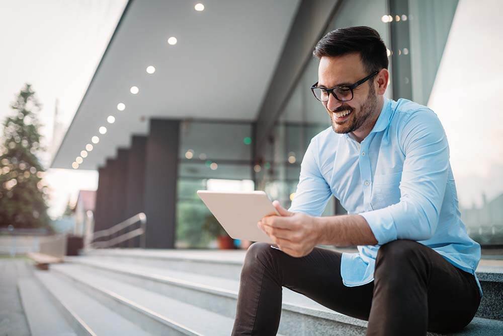 Selbstbewusster Geschäftsmann analysiert Immobilieninvestitionsdaten auf einem Tablet, während er auf den Stufen eines modernen Büros mit Glasarchitektur sitzt