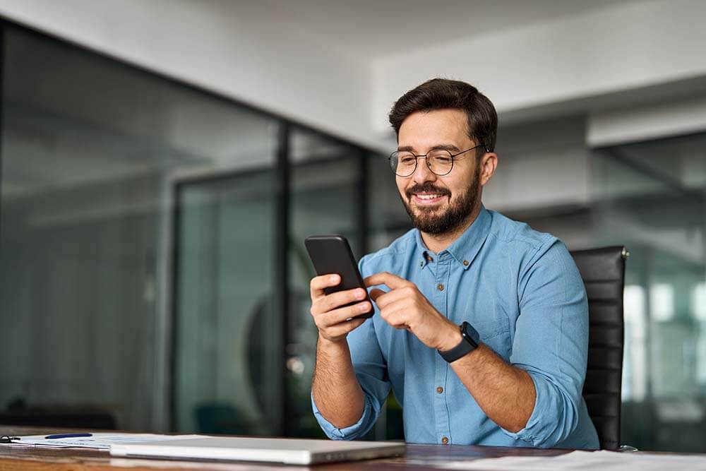 Lächelnder Eigentümer überprüft jährliche Steuerzahlungen für bulgarisches Eigentum auf dem Smartphone in einem modernen Glasbüro