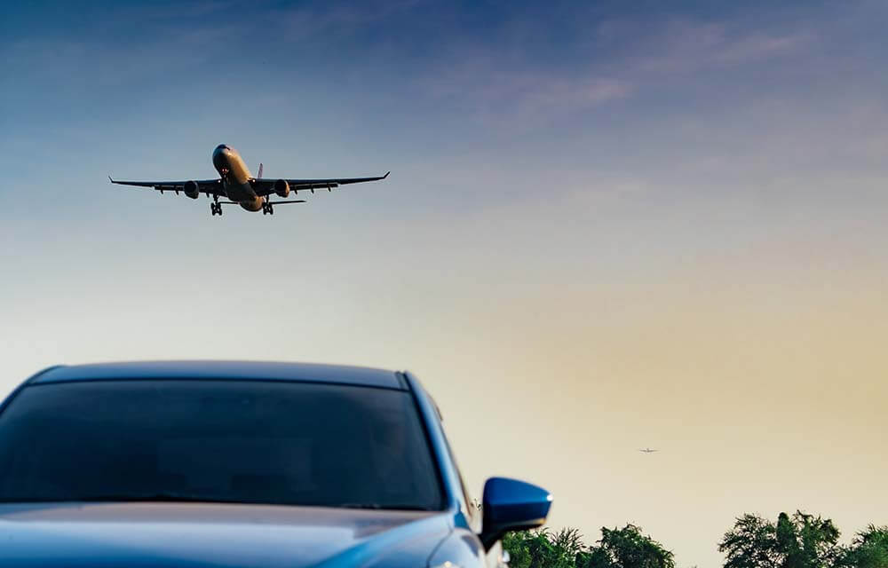 Ein Verkehrsflugzeug, das über die Motorhaube eines Luxusautos fliegt und den Luft- und Straßenverkehr vor blauem Himmel symbolisiert