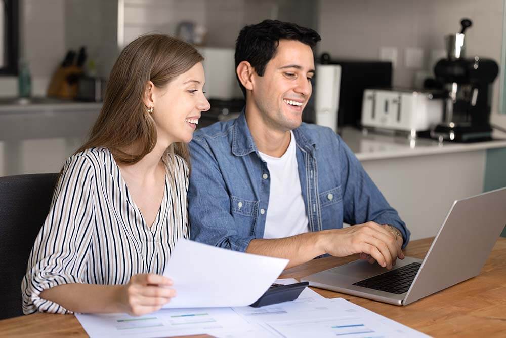 Glückliches Paar sitzt zusammen an einem Holztisch und studiert Dokumente zum Hauskauf auf einem Laptop, auf dem Papiere verteilt sind