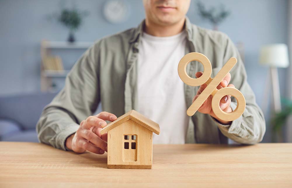 Erläuterung der Hypothekenbedingungen für finnische Immobilienkäufer Person mit Holzhausmodell und Prozentzeichen erklärt finnischen Immobilienkäufern die Hypothekenbedingungen