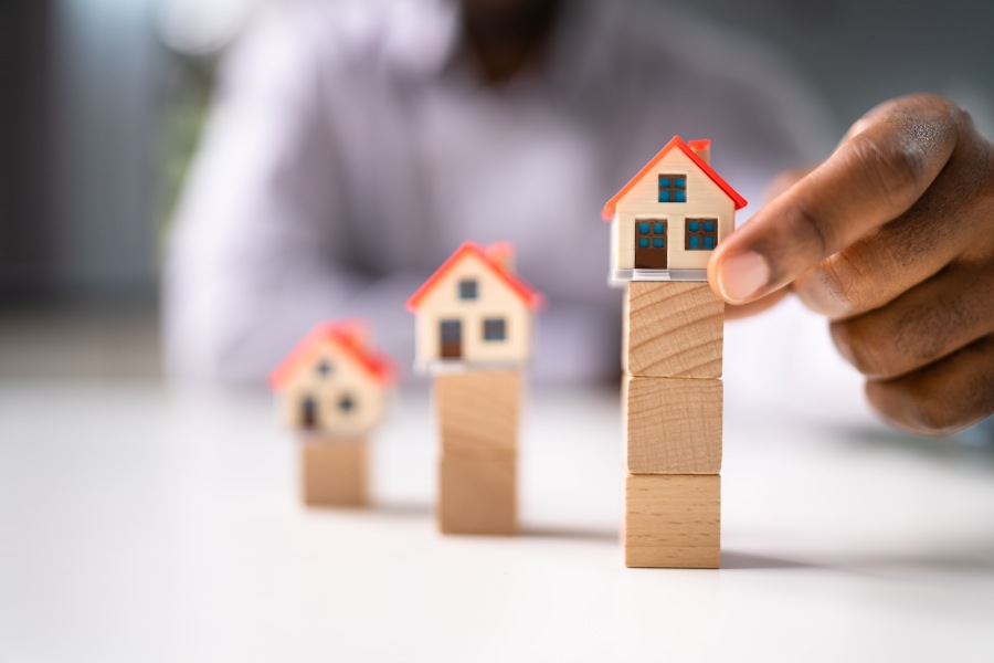 Eine Hand ordnet Hausmodelle auf Holzblöcken an und zeigt Preisabstufungen sowie Risiken des Rohbaumarktes in Bulgarien.